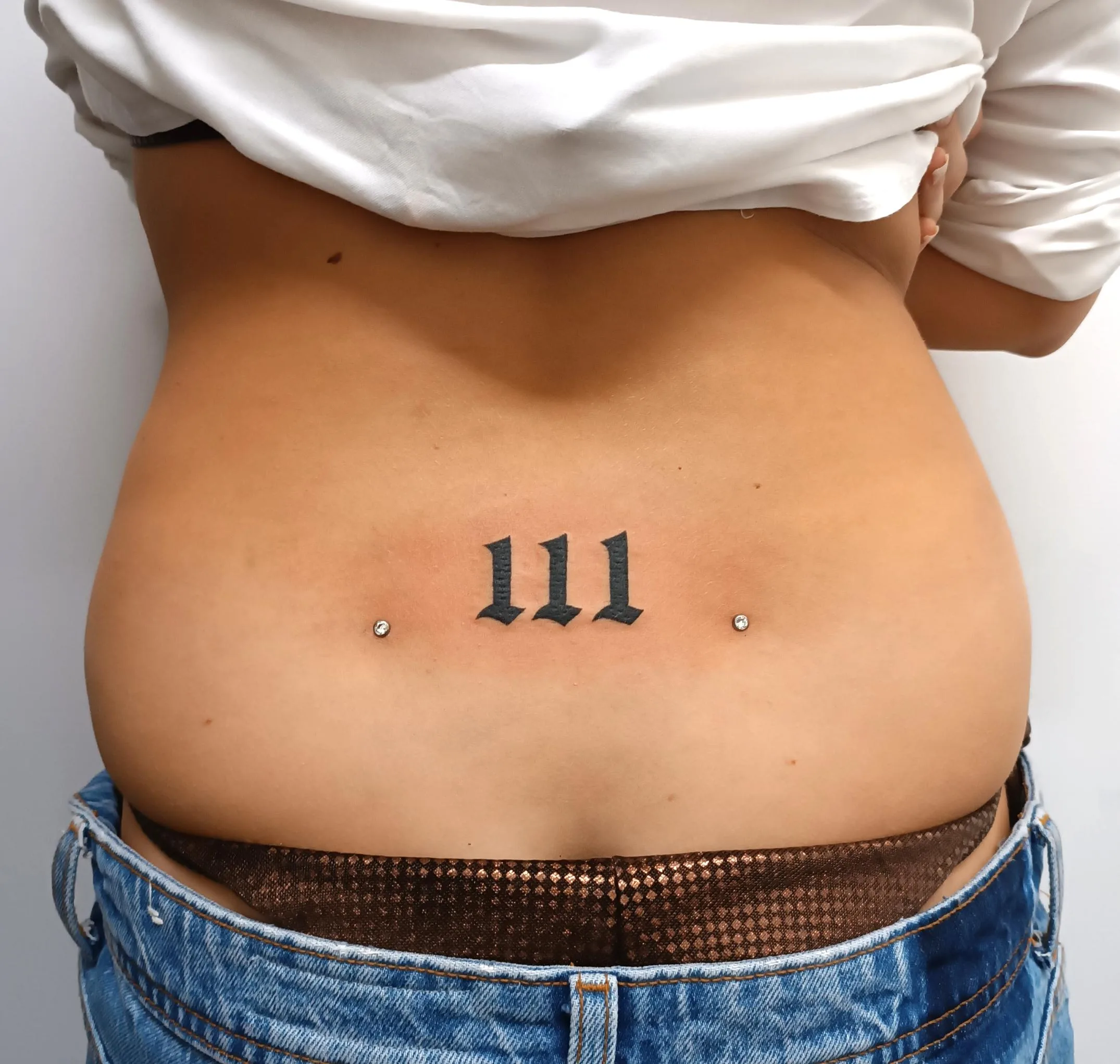 Tatuaje en la espalda baja con "111" en gótico negro, flanqueado por dos microdermales brillantes.