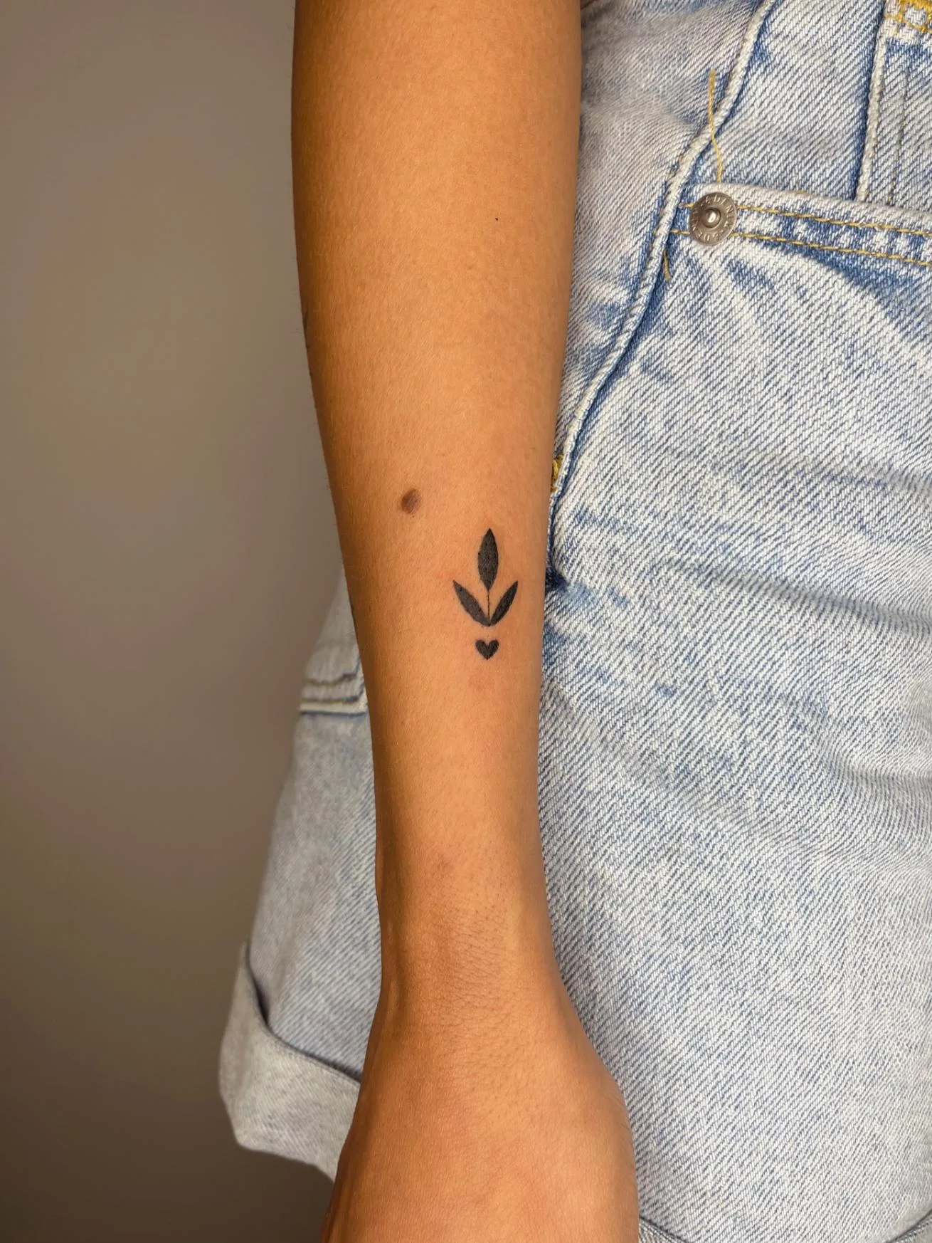 Tatuaje en el brazo de una rama curvada con hojas negras y un mini corazón negro.