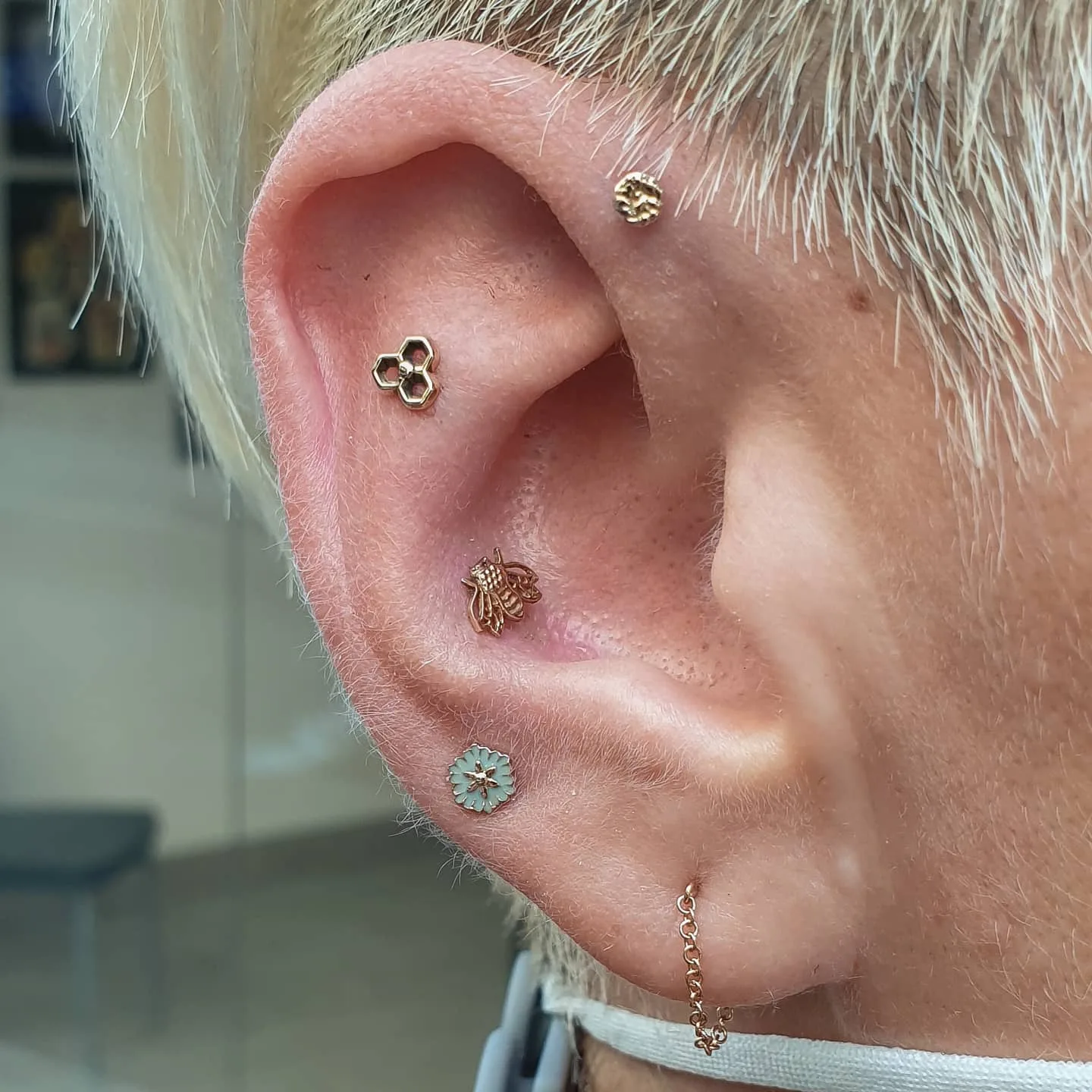 Vista de una oreja con varios piercings decorados con joyería de oro, incluyendo una pieza con diseño de abeja, un hexágono y un pendiente colgante en la parte inferior.