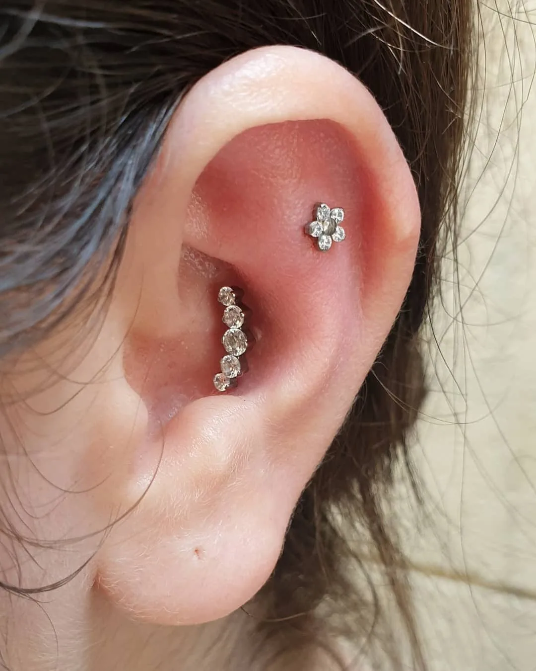 magen de una oreja con un piercing en la concha adornado con una línea de cristales, acompañado por un pendiente de flor en el lóbulo.