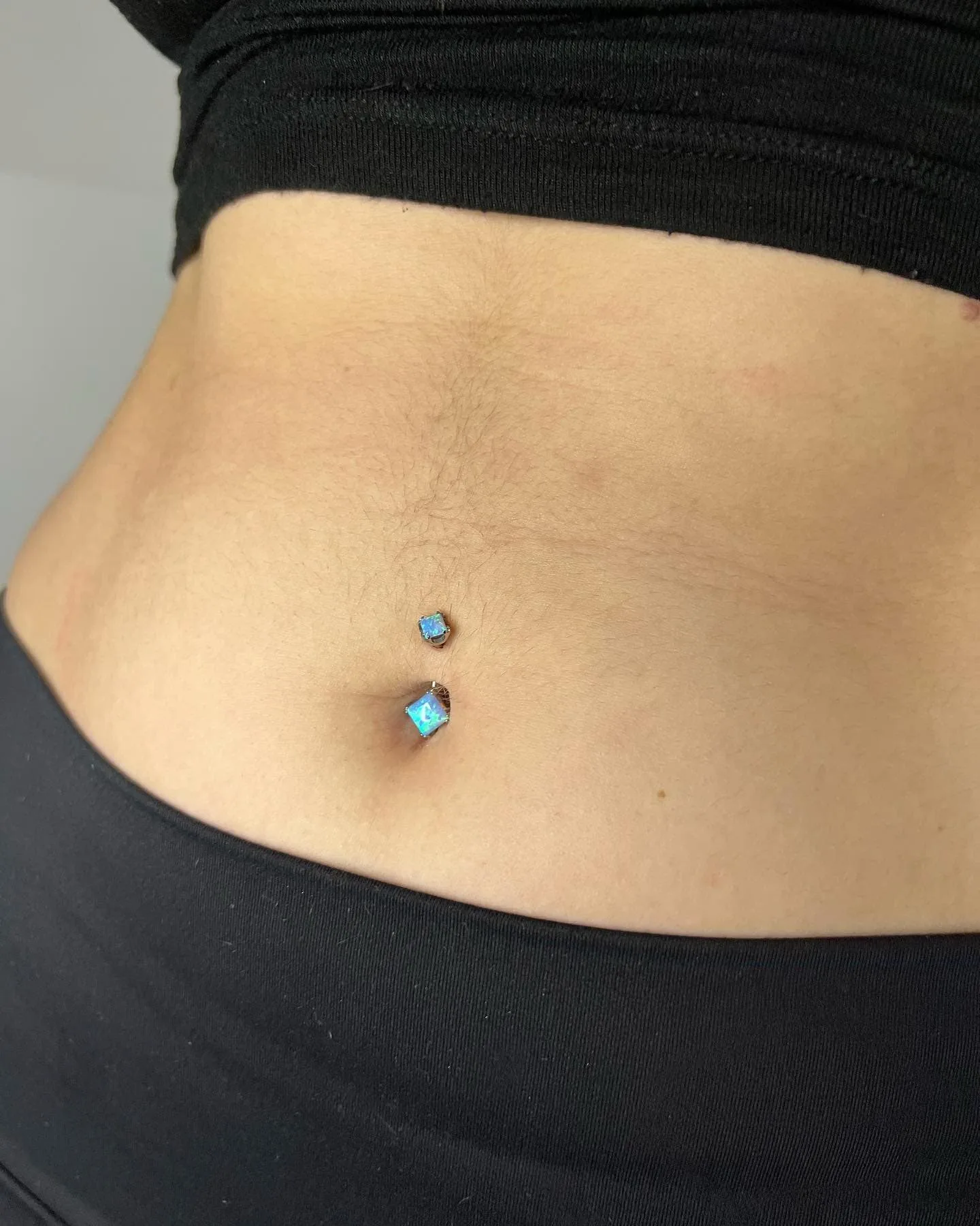 Piercing de ombligo doble con joyería de ópalo azul, ubicado justo encima del ombligo y en la piel superior del abdomen.