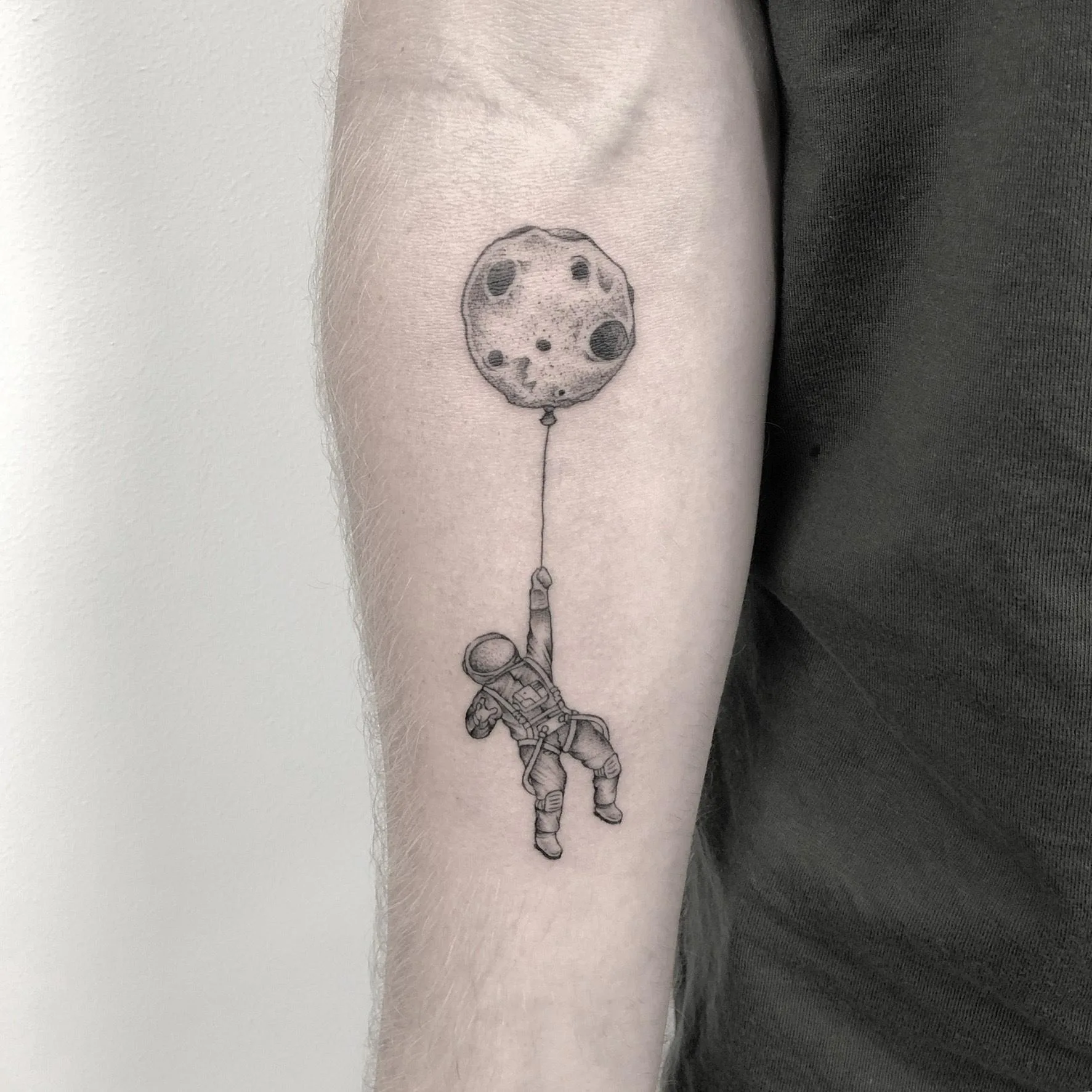 Tatuaje de un astronauta sujetando una luna en forma de globo en el antebrazo, estilo microrealismo en tonos de gris.