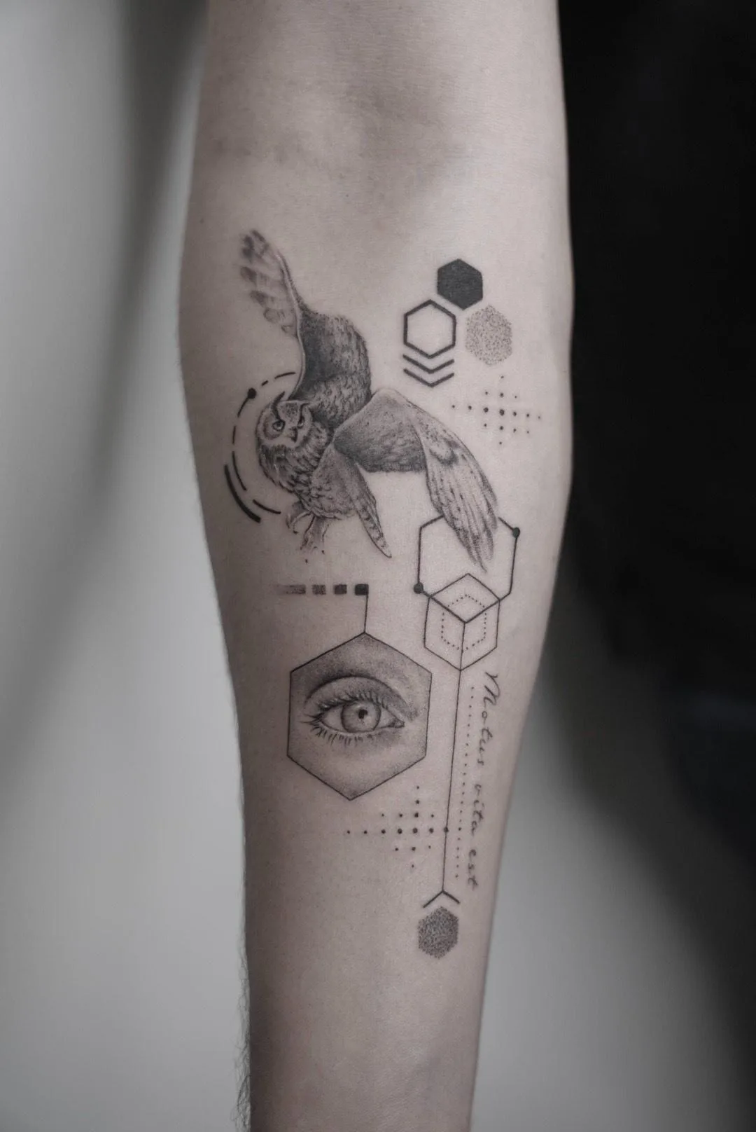 Tatuaje en el antebrazo de un búho en vuelo, rodeado de figuras geométricas y un ojo en estilo microrealismo.