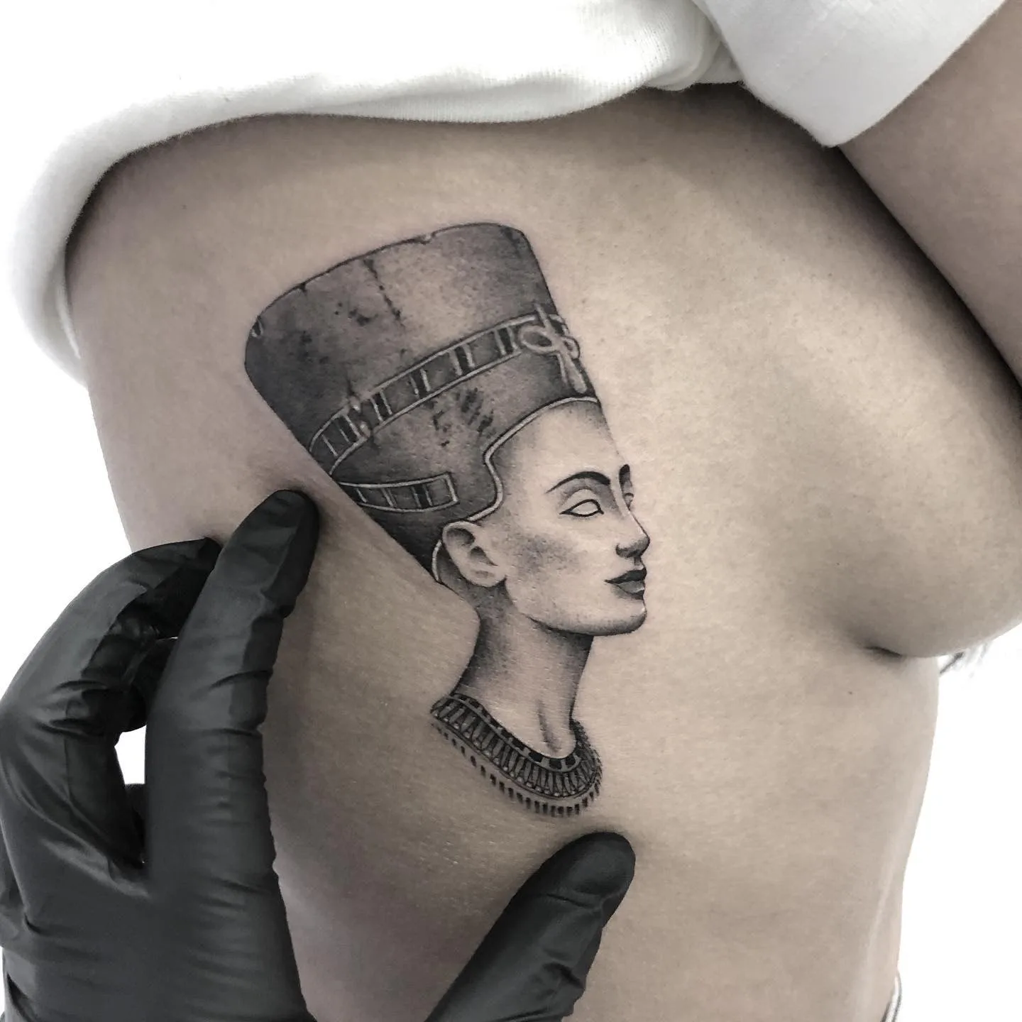 Tatuaje de la reina Nefertiti en el costado de una persona, estilo microrealismo en tonos grises y negros.