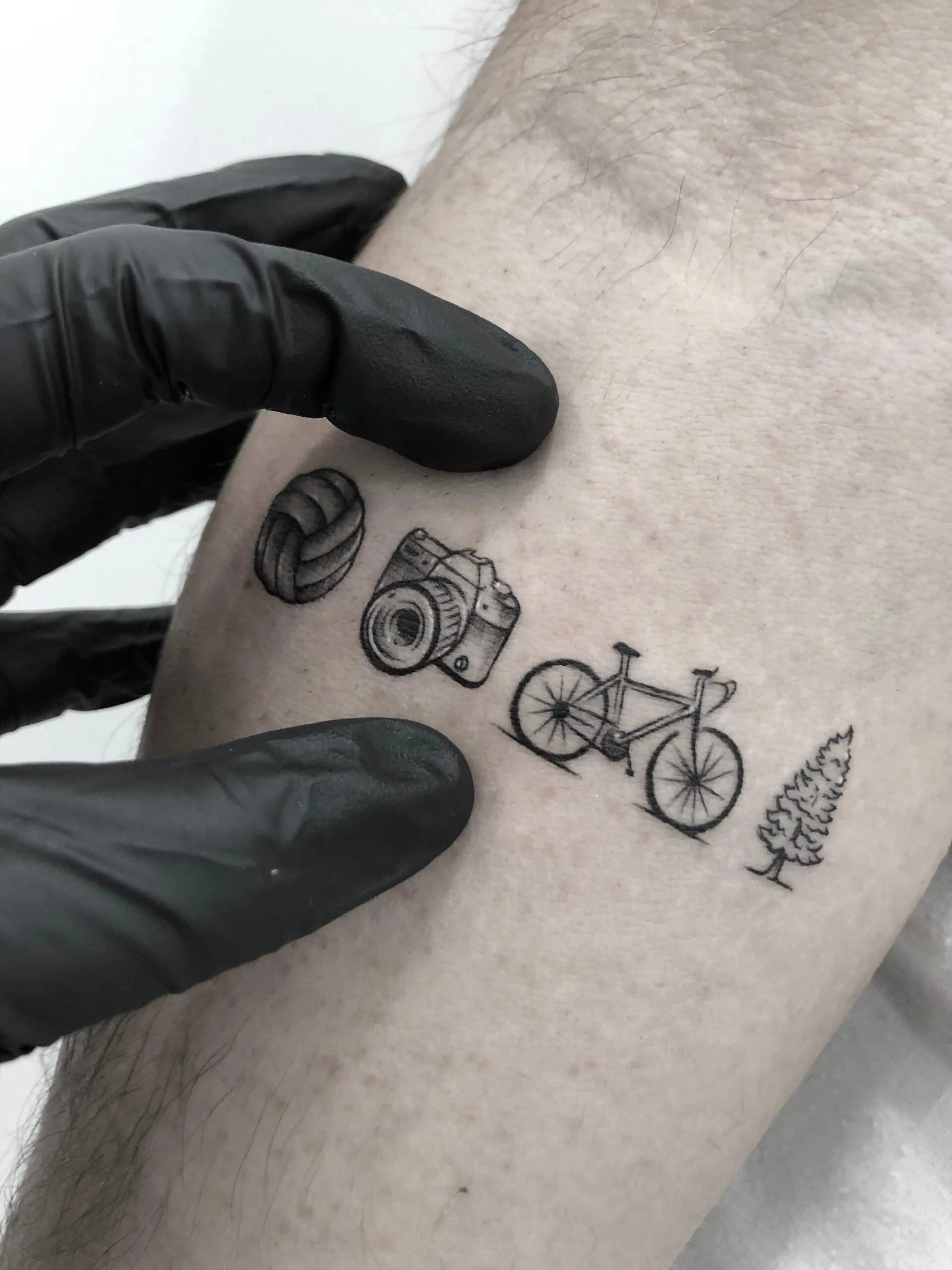 Tatuaje de estilo minimalista en el brazo, con iconos de una pelota, una cámara, una bicicleta y un árbol en tinta negra.