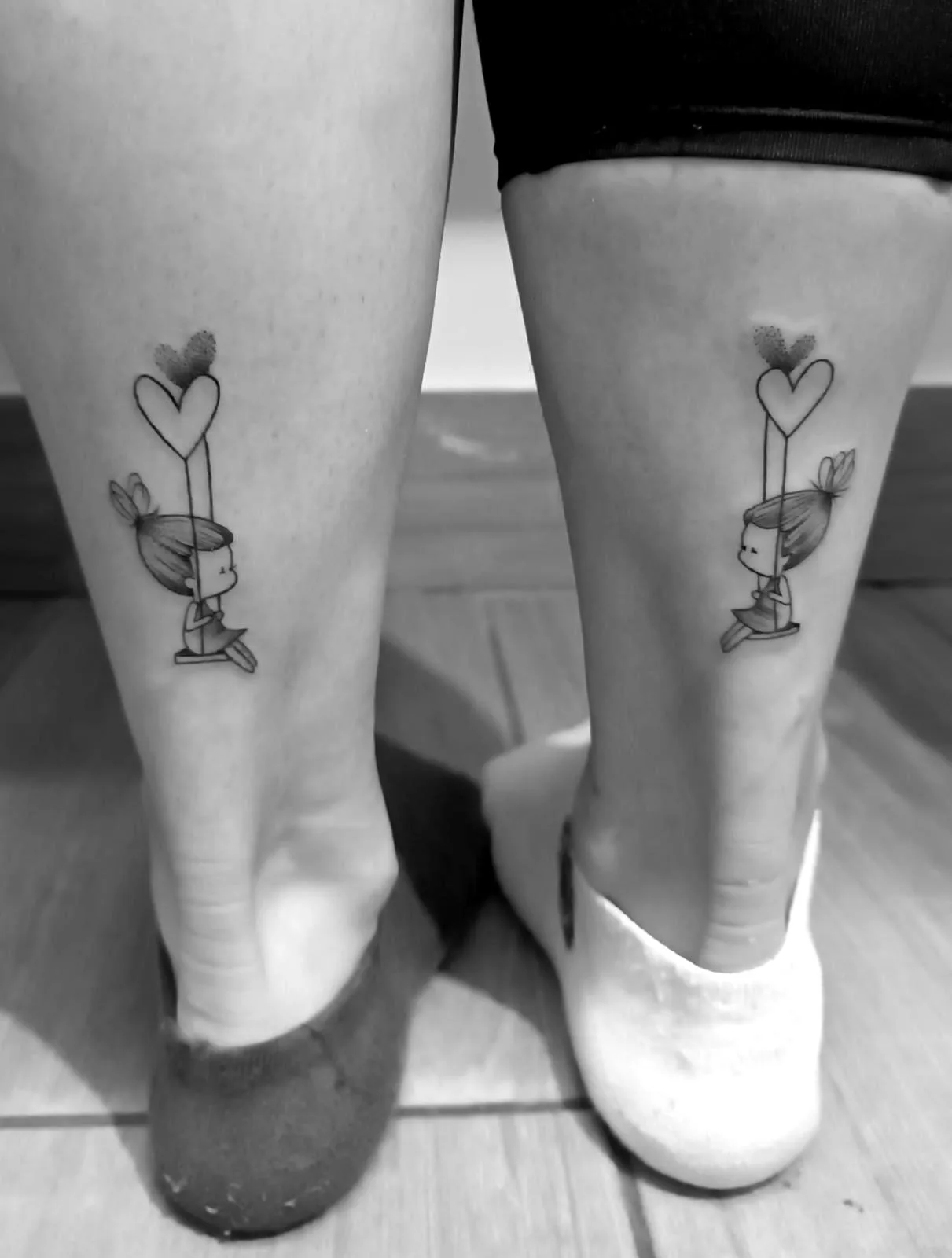 Tatuaje de pareja en las piernas, con un diseño de niña sentada en un columpio colgado de un corazón.