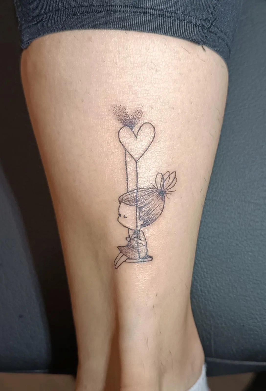 Tatuaje de una niña en columpio, estilo minimalista en la pierna, colgando de un corazón.