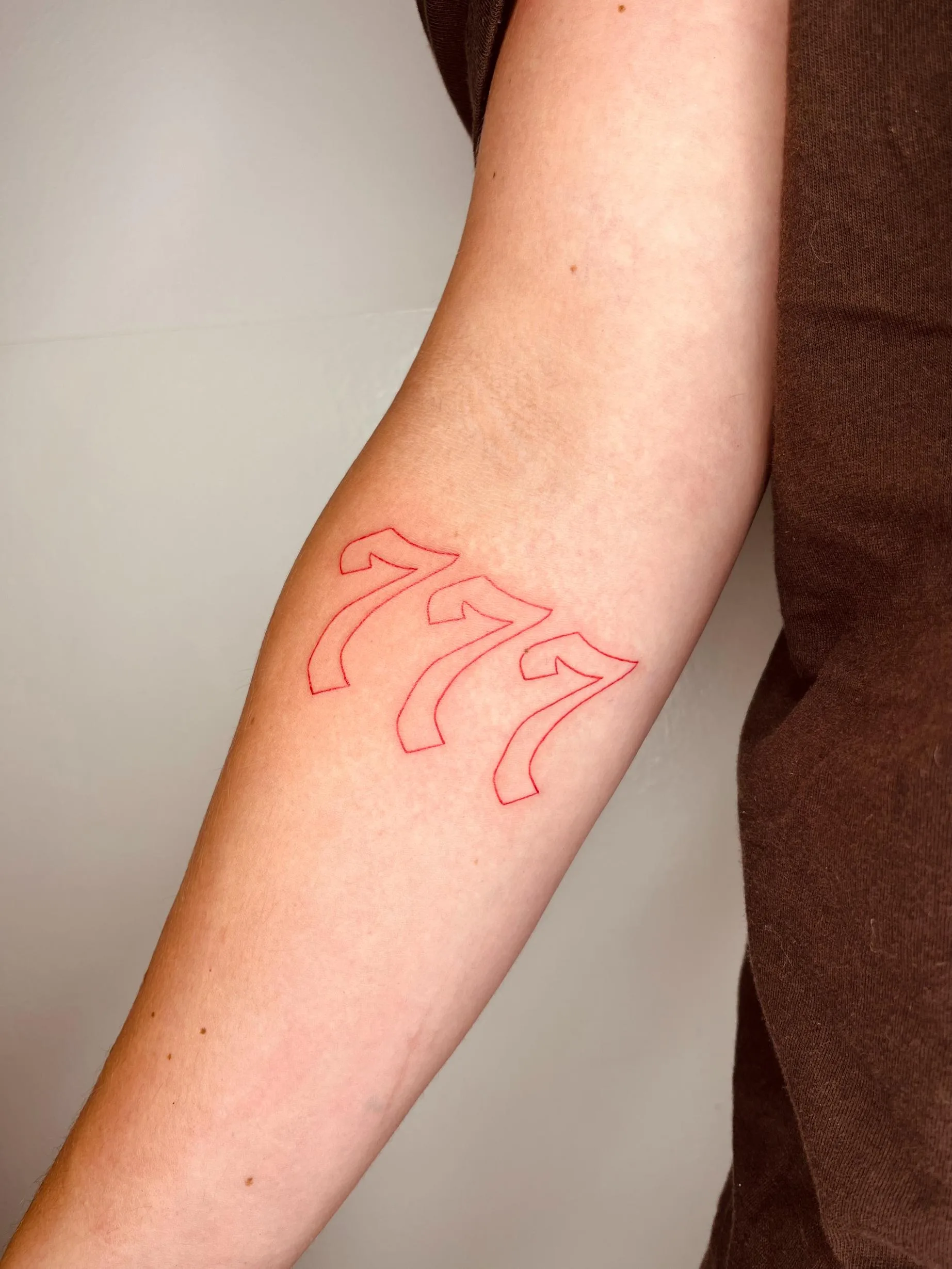 Tatuaje de números "777" en el antebrazo, realizados en tinta roja con un estilo de contorno simple.