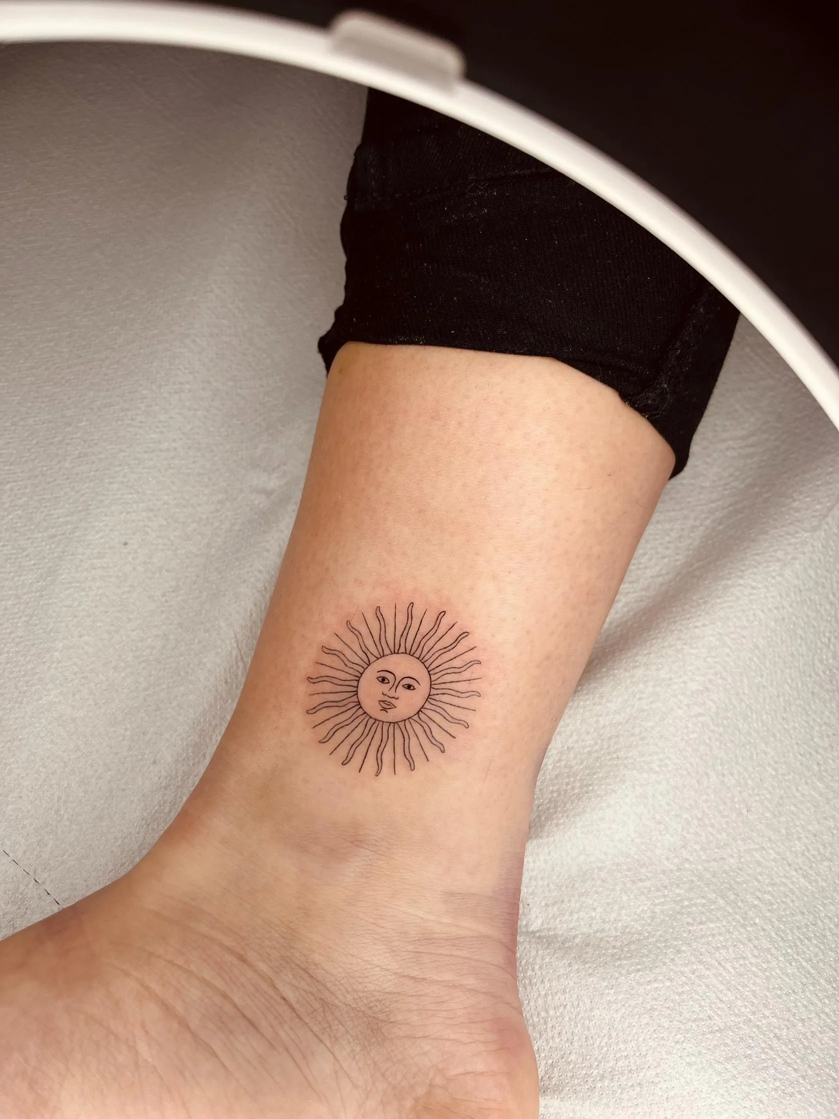 Tatuaje minimalista de un sol con rostro en el tobillo, con rayos del sol en líneas finas.
