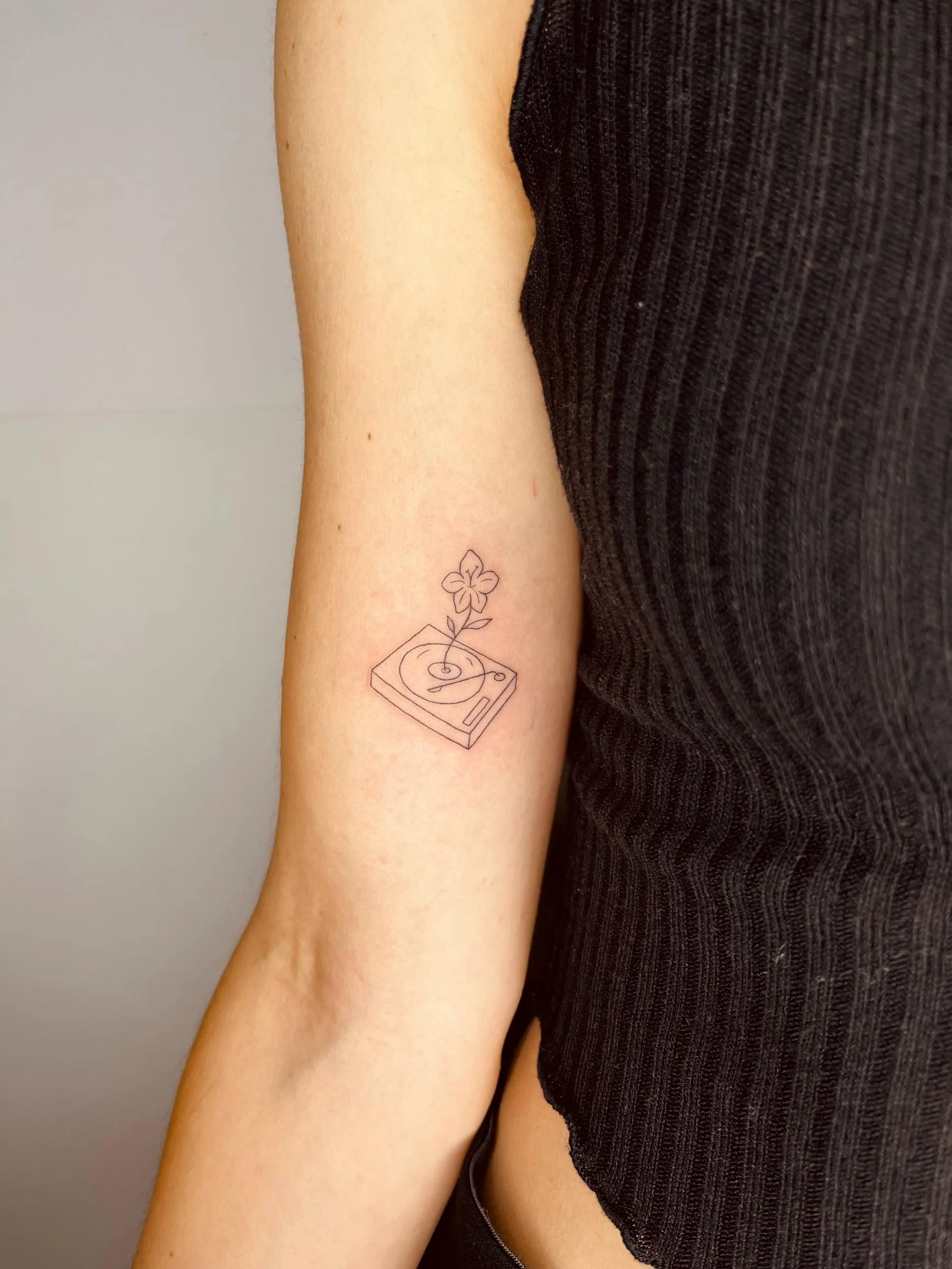 Tatuaje de estilo minimalista en el brazo, con un diseño de tocadiscos y un pequeño trébol sobre él, líneas finas y simples.