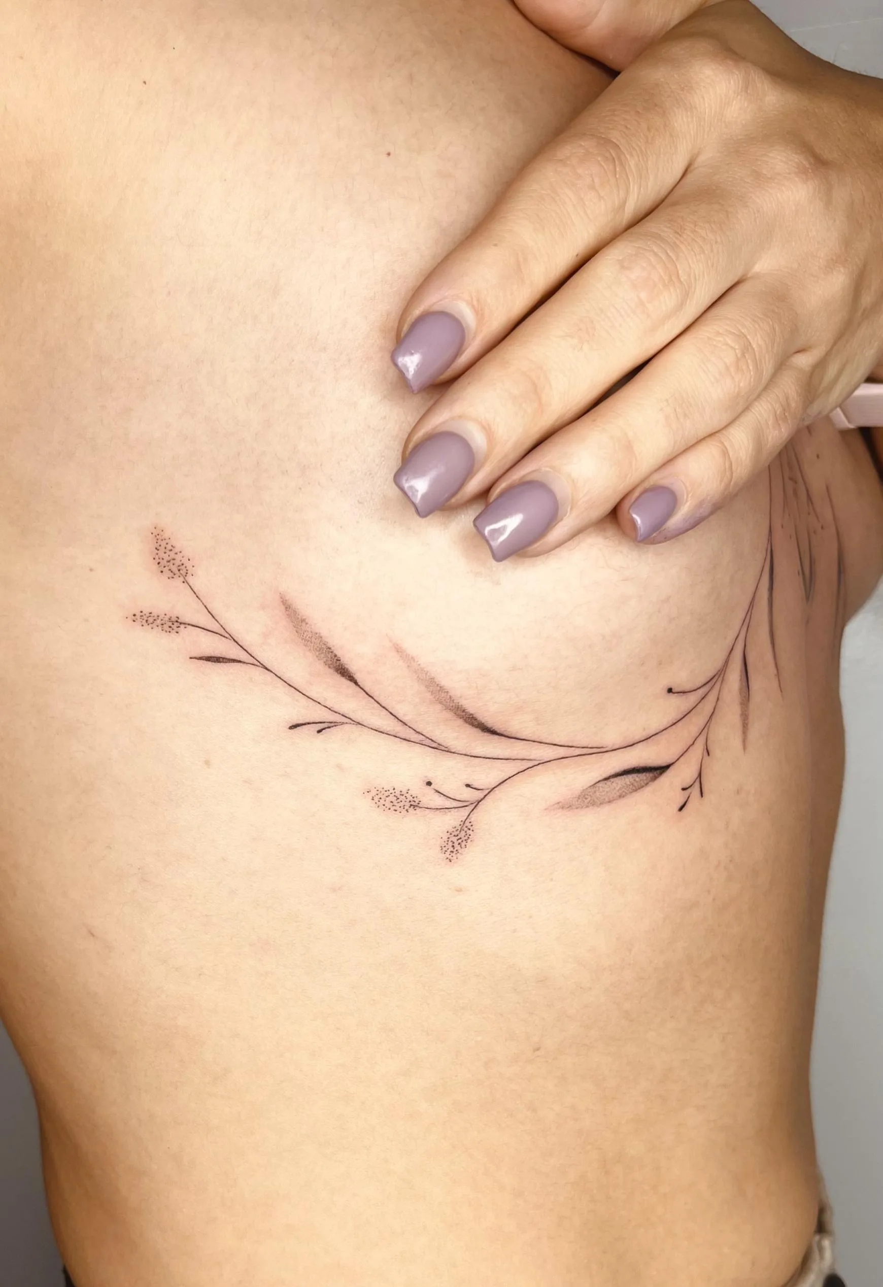 Tatuaje minimalista de una ramita con pequeñas hojas y semillas debajo de los senos, realizado en tonos delicados y con puntillismo en algunos detalles,